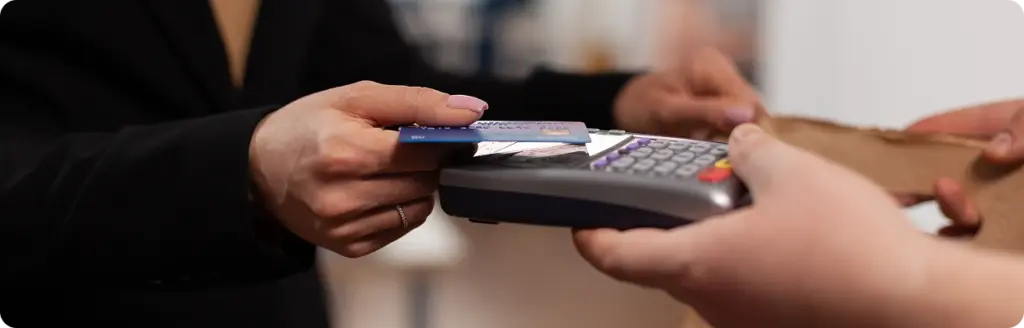 Een persoon in een zwart pak voert een contactloze betaling uit met een kaart bij een kaartlezer. In de andere hand houdt hij een papieren tas vast, wat duidt op een aankoop