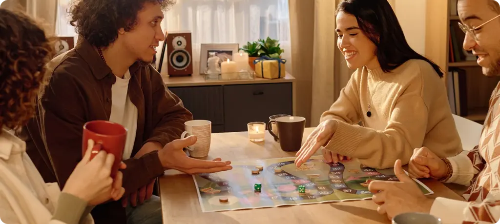 Vier vrienden spelen een bordspel aan een gezellige tafel, bij sfeervol licht en met een drankje erbij. De sfeer is vriendelijk en uitnodigend, waarbij de nadruk ligt op gezellig samenzijn